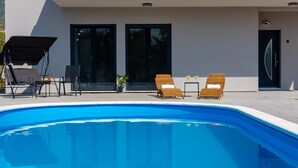 Ein moderner Poolbereich mit klarem, blauem Pool, zwei Holzliegen, einem kleinen Tisch mit Topfpflanze und einer überdachten Sitzgruppe.