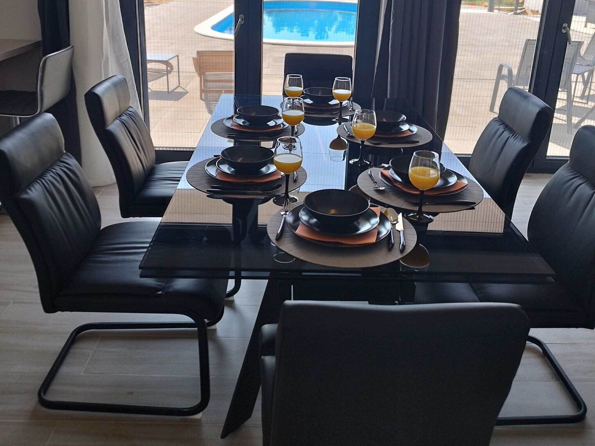 A dining area with a pool view, black dining chairs, and a table set for four with orange juice glasses.