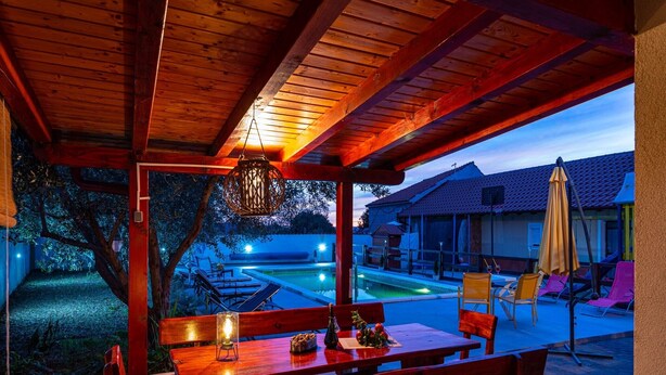An outdoor patio with wooden tables and benches, a hanging light fixture, and a view of a swimming pool and houses in the background.