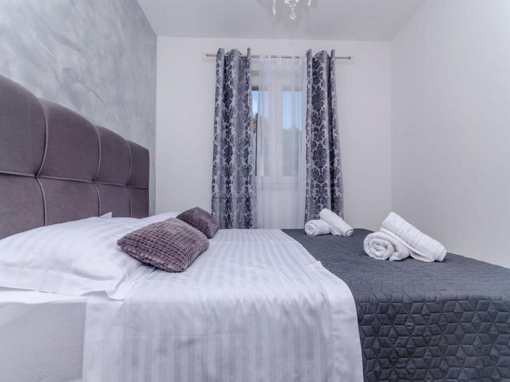 Un dormitorio moderno con una cama grande, dos almohadas, una mesita de noche con toallas dobladas y una ventana con cortinas estampadas.