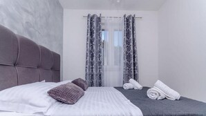 Une chambre moderne avec un grand lit, deux oreillers, une table de chevet sur laquelle sont pliées des serviettes et une fenêtre agrémentée de rideaux à motifs.