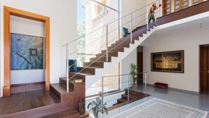 Un intérieur spacieux avec un escalier menant à un niveau supérieur, une grande peinture abstraite sur le mur de gauche et une grande peinture de paysage sur le mur de droite.