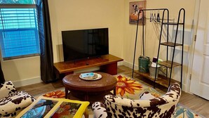 A living room with a television, a wooden coffee table, two cow-patterned armchairs, a colorful rug, and a metal shelf unit.