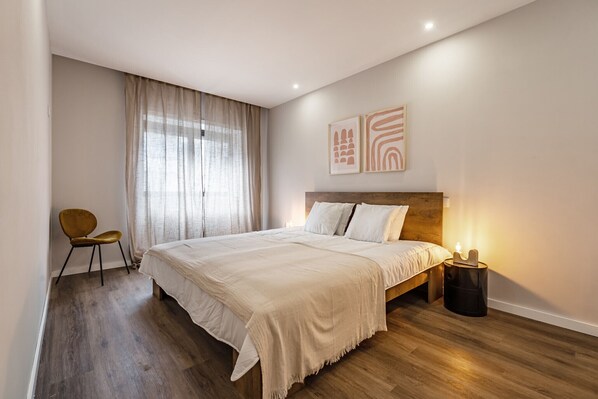 A modern bedroom with a large bed, a wooden floor, a chair, and a bedside table with a lamp.