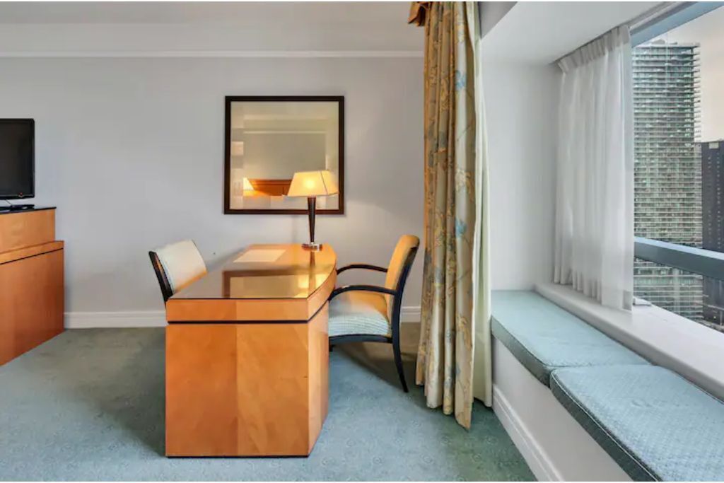 Ett hotellrum med ett skrivbord, en stol, en lampa och ett stort fönster med gardiner.