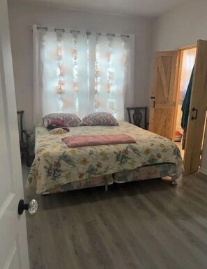 Une chambre à coucher avec un lit, une literie à motifs floraux, une porte en bois et une fenêtre avec des rideaux.