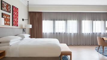 A modern hotel room with a large bed, a desk, and a chair. There are curtains on the windows and a lamp on the wall.