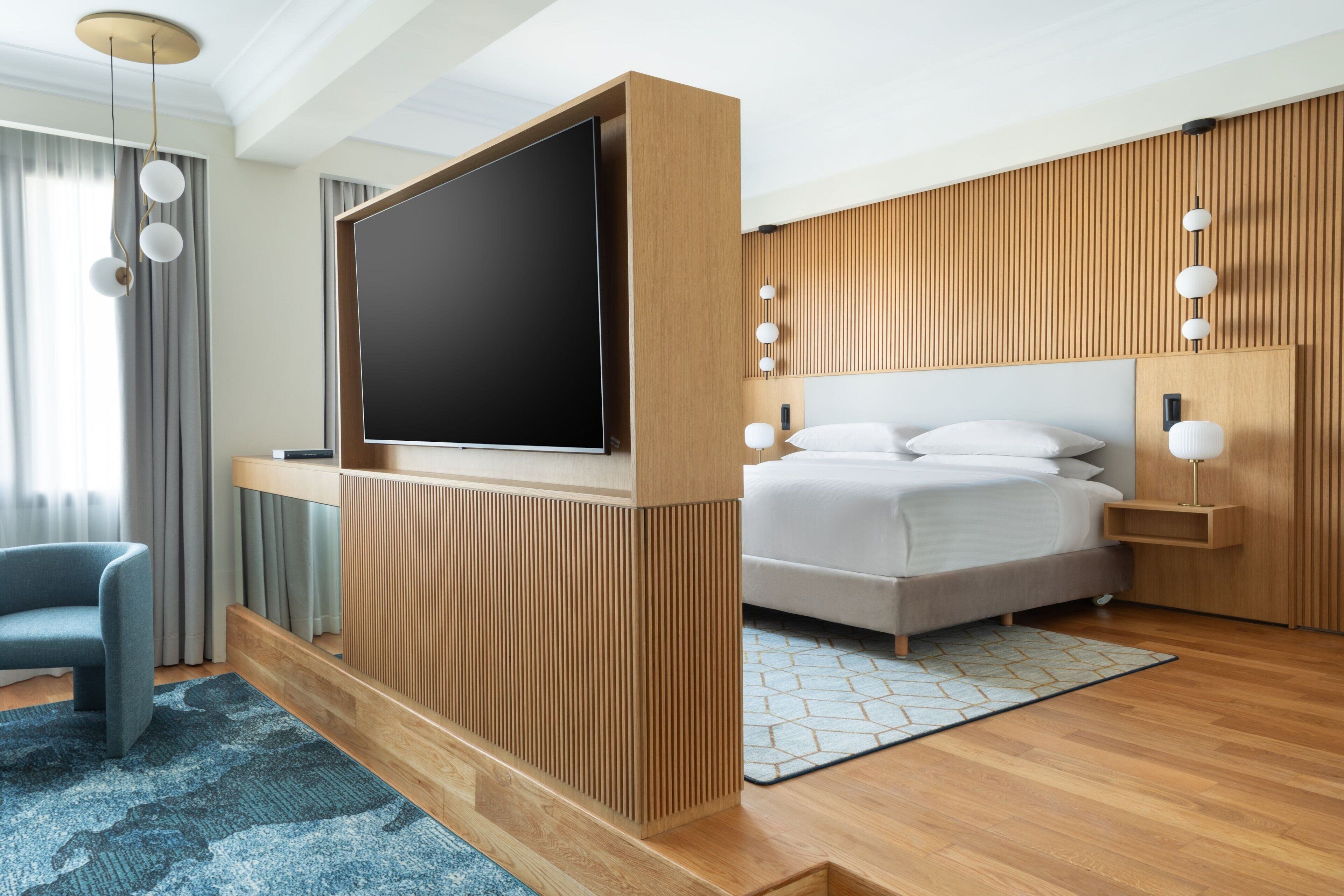 Quarto de hotel moderno com uma televisão de tela plana grande, uma cama com roupa de cama branca, um tapete azul estampado e painéis de madeira nas paredes.