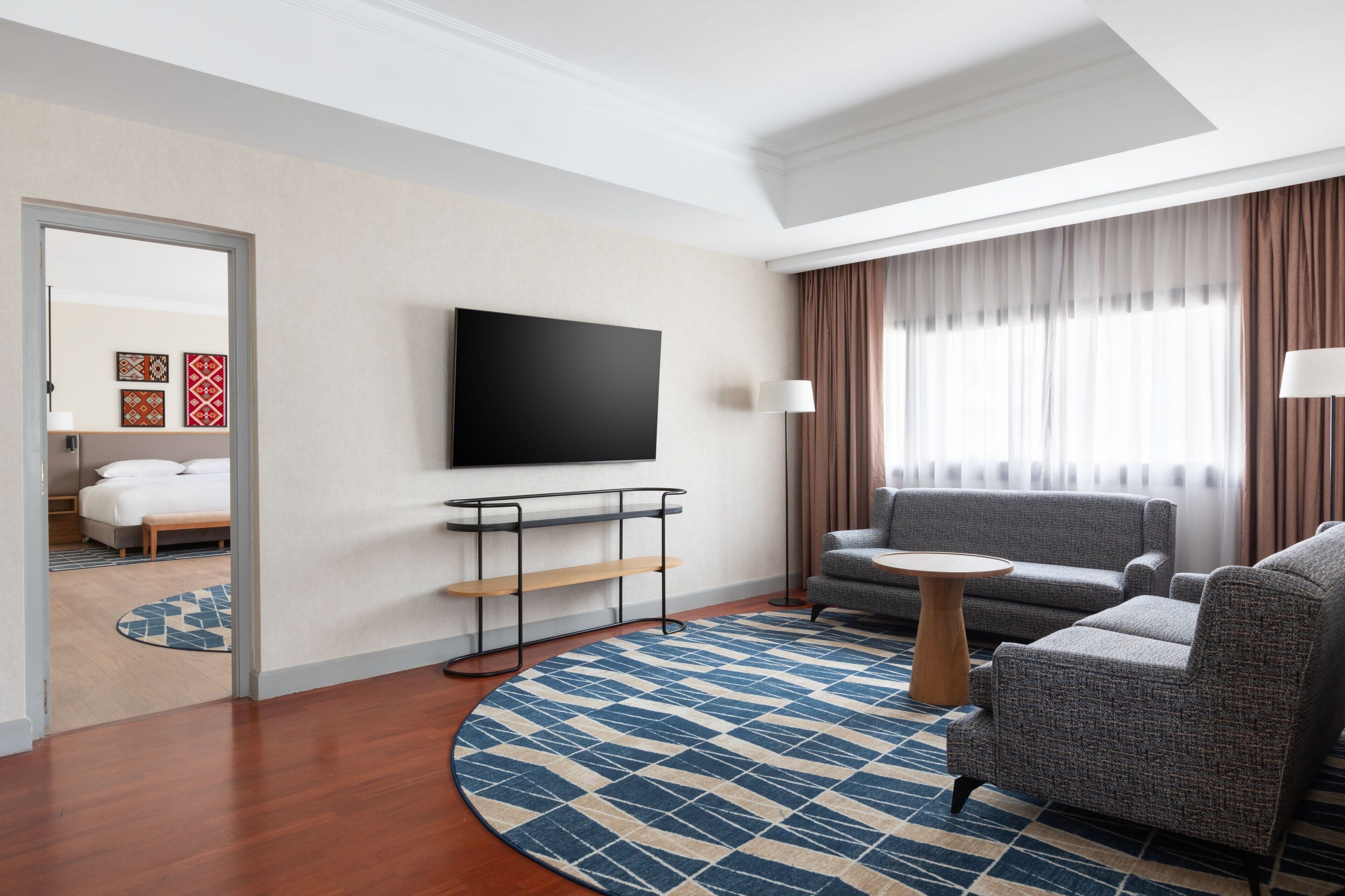 A modern hotel room with a flat-screen TV mounted on the wall, a sofa, a small round table, and a patterned rug.