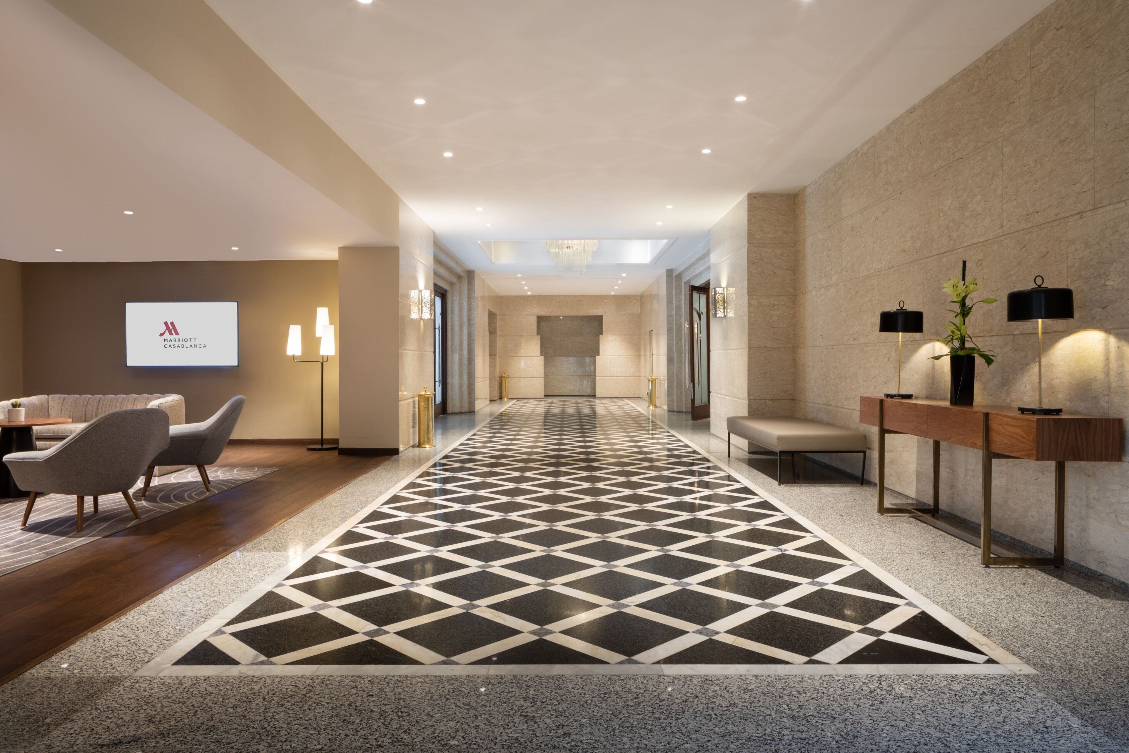A modern hotel lobby with a geometric patterned floor, a reception desk, seating area, and a projection screen displaying the hotel name.