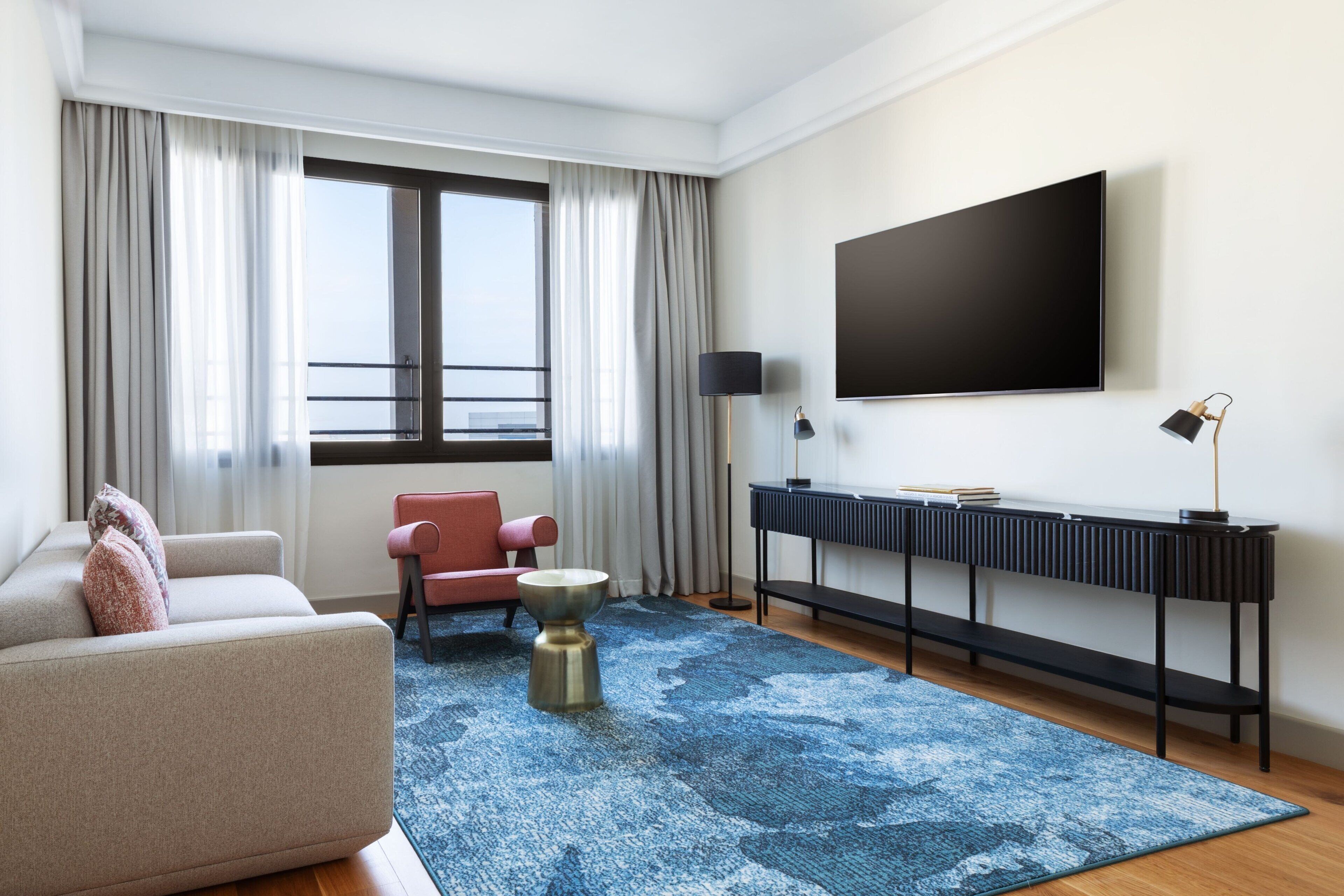 A modern hotel room with a large window, a flat-screen TV, a sofa, a chair, a coffee table, and a console table with a lamp.