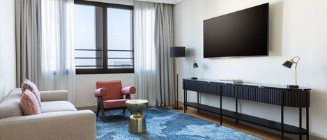 A modern hotel room with a large window, a flat-screen TV, a sofa, a chair, a coffee table, and a console table with a lamp.