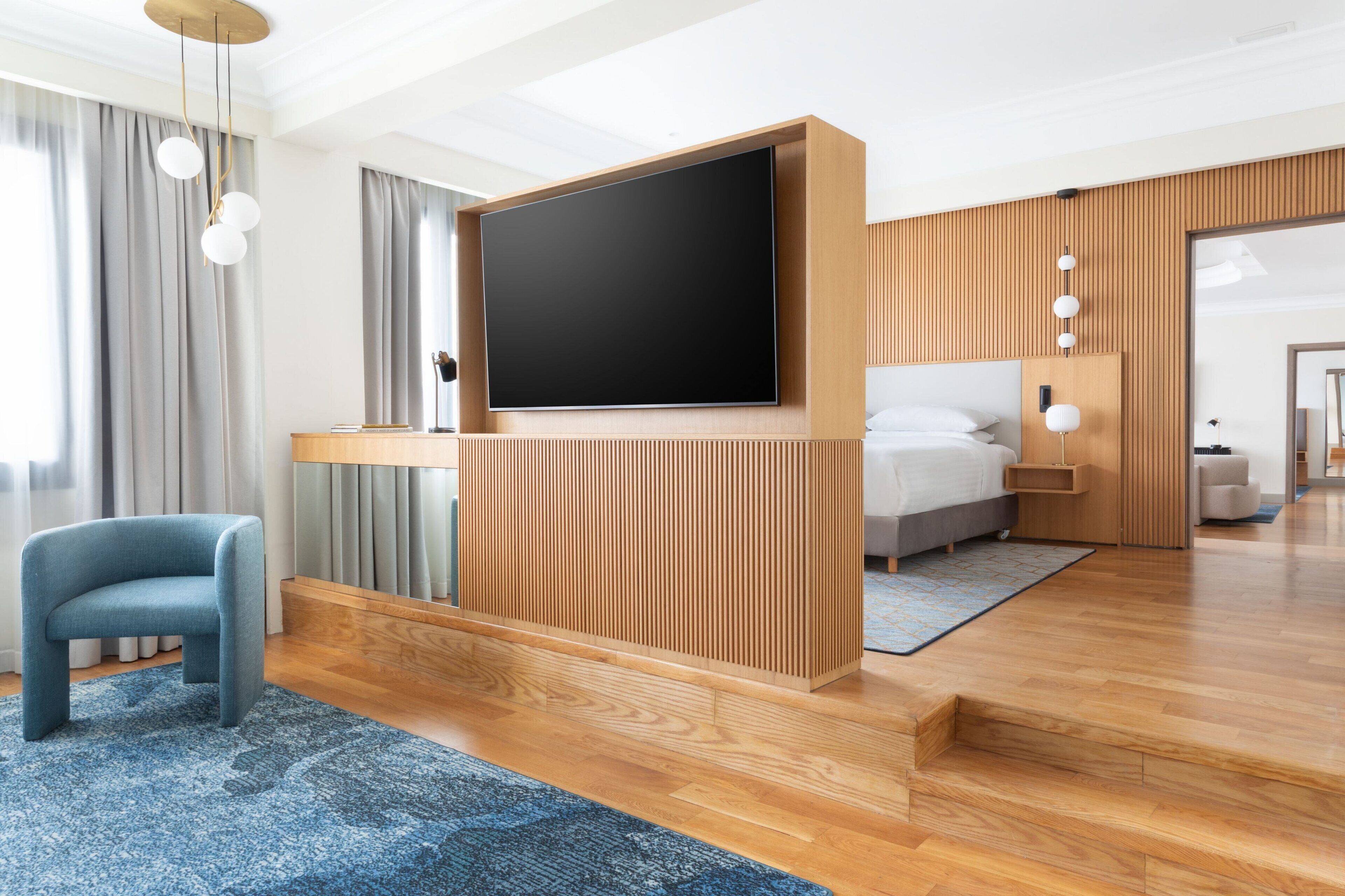 A modern hotel room with a large flat-screen TV, a wooden accent wall, a bed, a desk, and a blue chair.