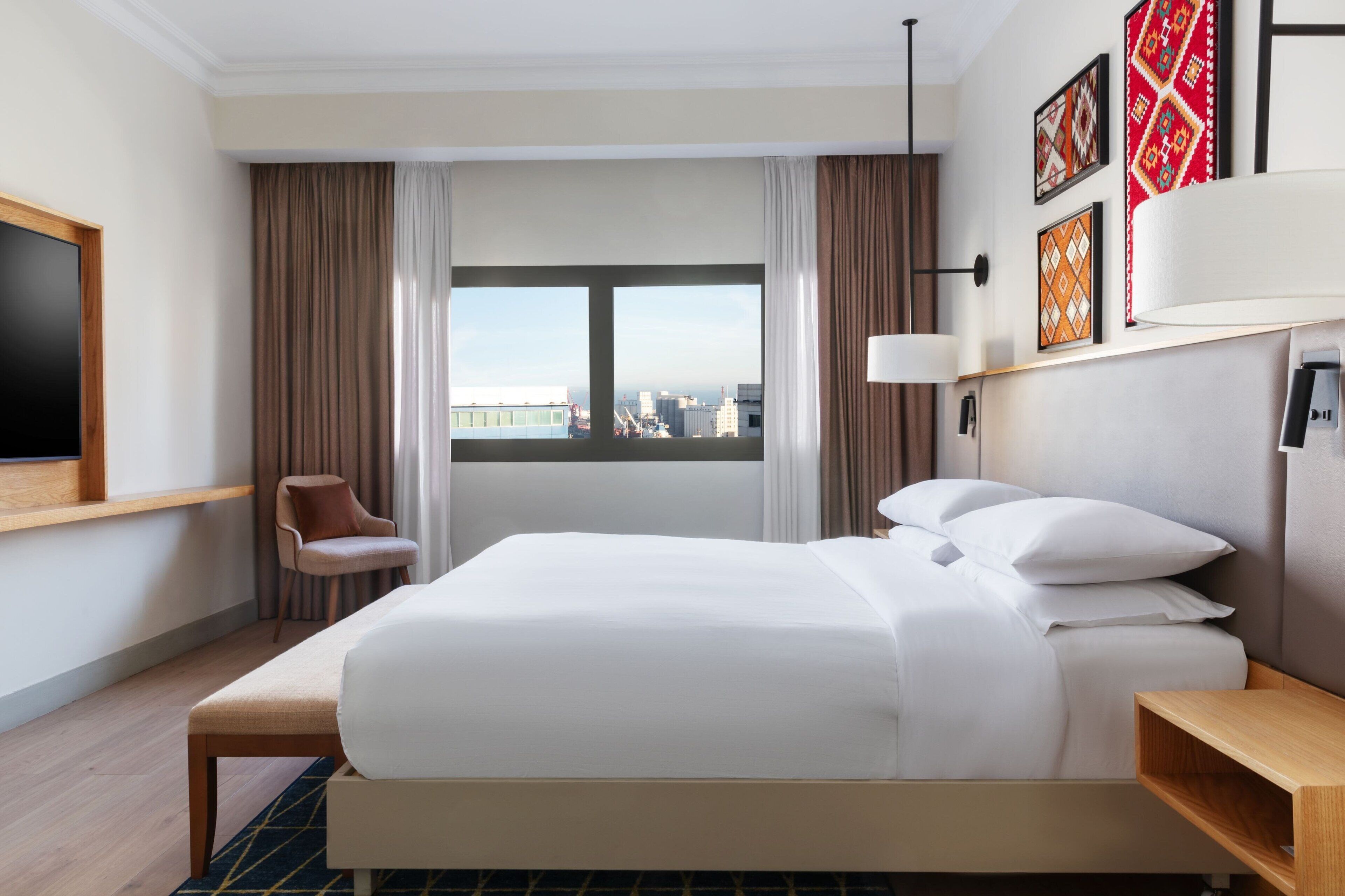 A hotel room with a large bed, a chair, a TV, and a window with a city view.