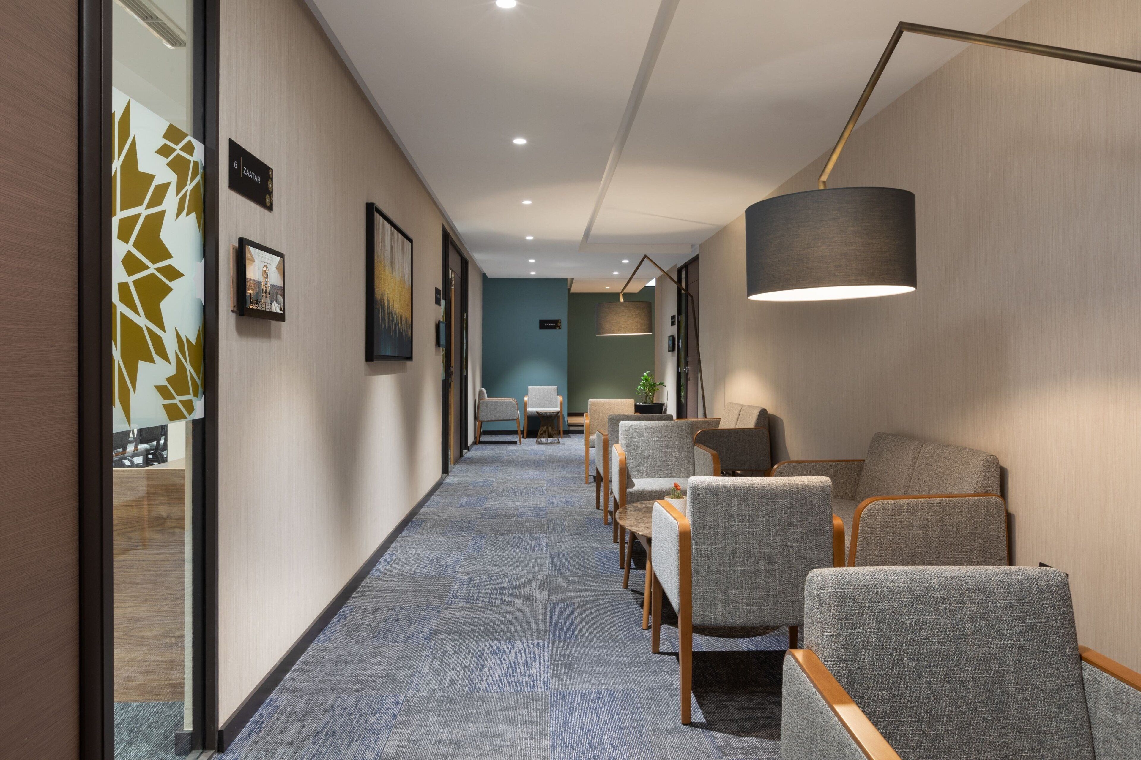A hotel corridor with seating areas, framed artwork, and a lamp.