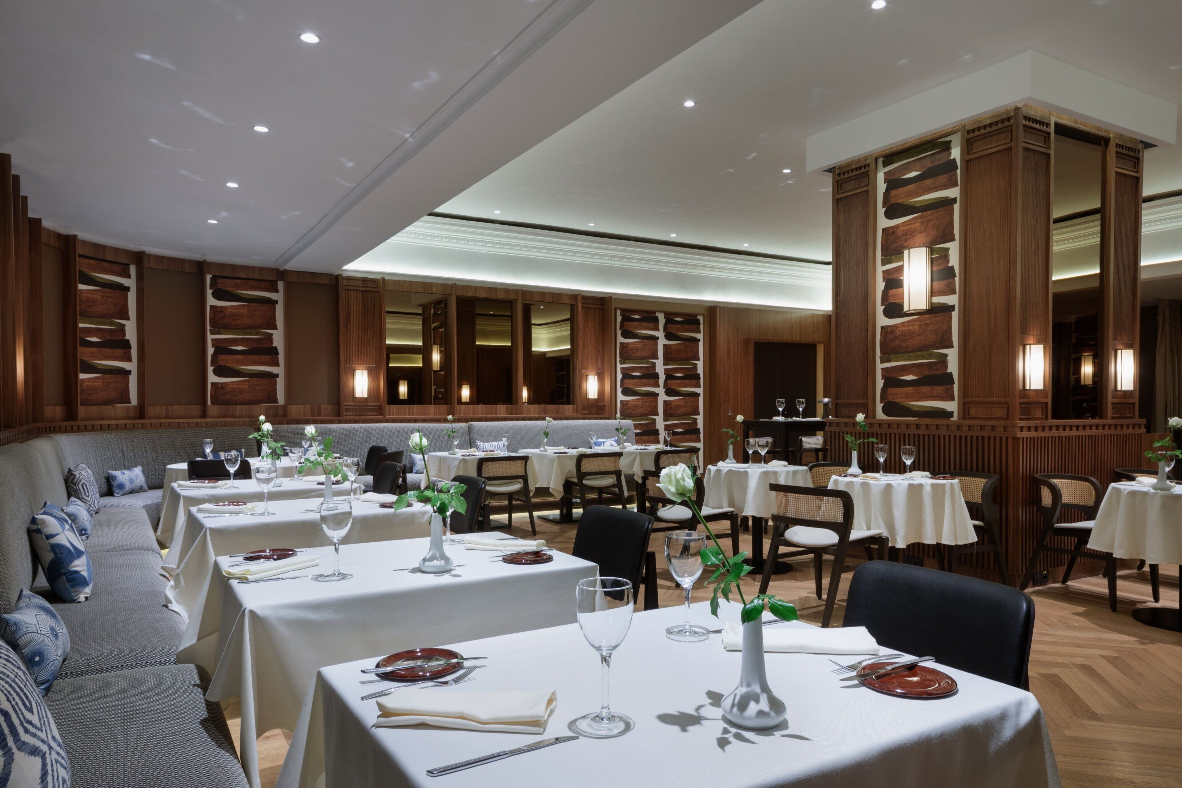 Un restaurant doté de nombreuses tables, chacune recouverte d’une nappe blanche, agrémentée de verres à vin et de couverts. Des coins salons confortables sont aménagés le long des murs, et le plafond est haut, avec des luminaires encastrés.