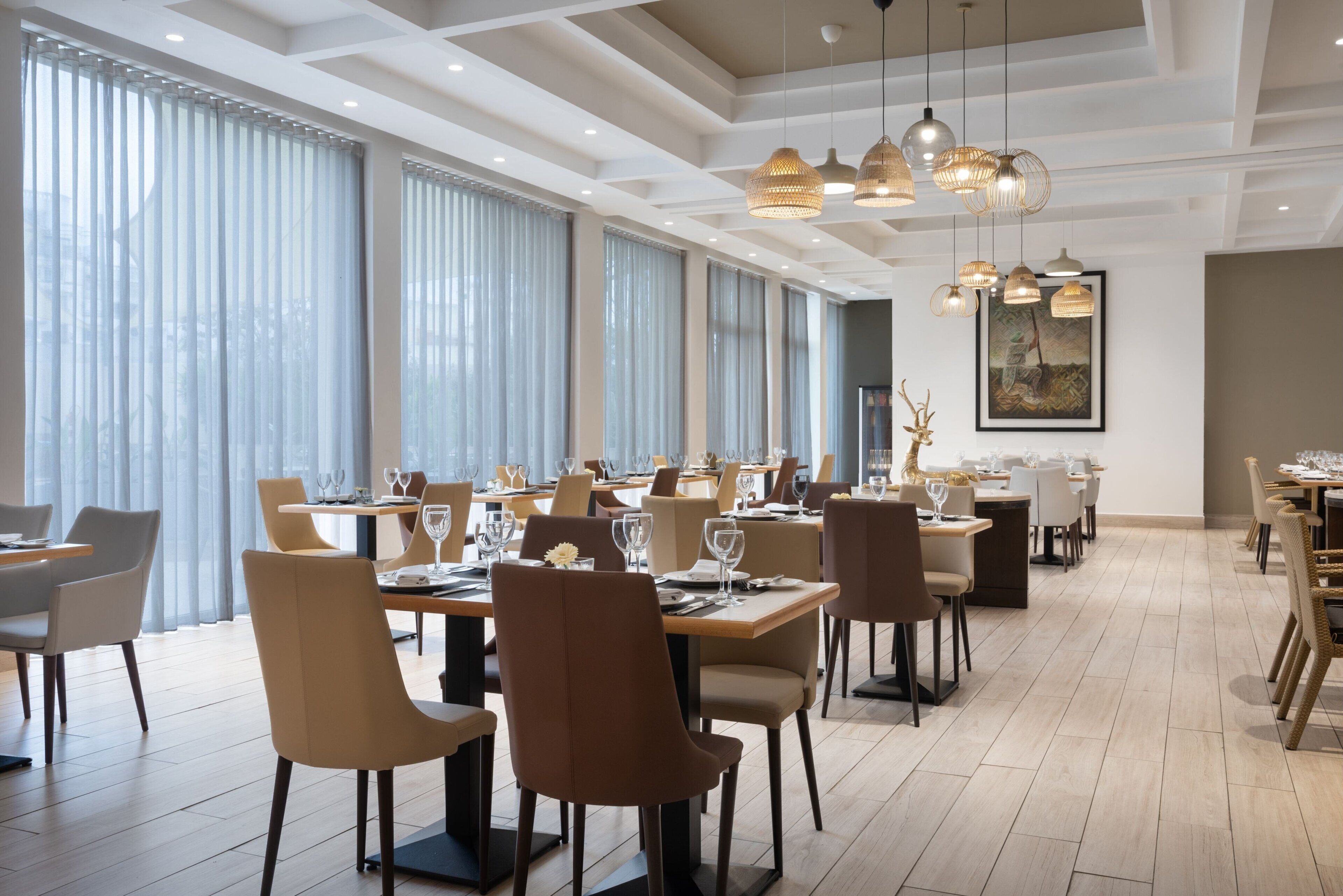Un comedor amplio con varias mesas y sillas, ventanas grandes con cortinas y lámparas colgantes.