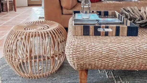 Una sala de estar acogedora con un sofá marrón, un reposapiés tejido y una mesa de centro con libros y objetos decorativos.