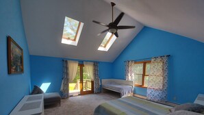 Un dormitorio con techo inclinado, tragaluces, ventilador de techo, cama con manta a rayas, una banca y un escritorio.