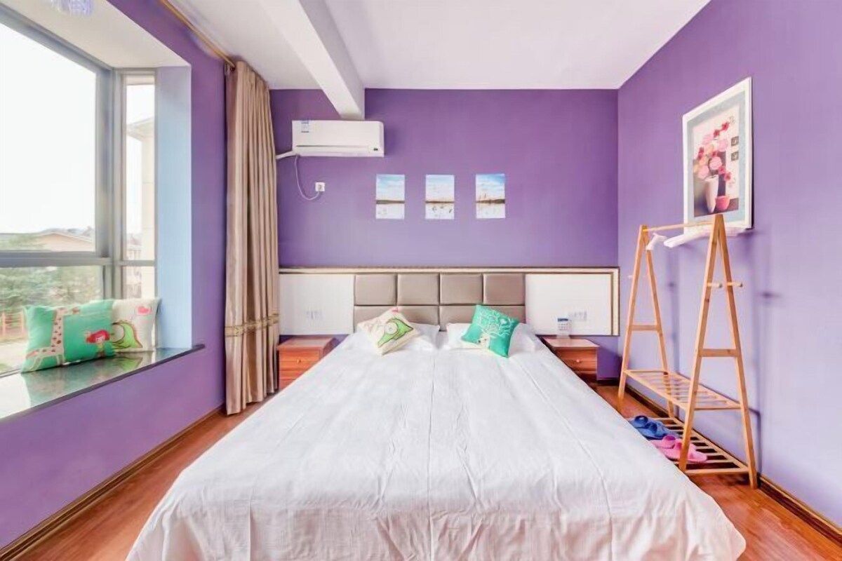 A bedroom with a large bed, a window with curtains, a wooden ladder, and a wall-mounted air conditioner.