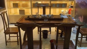 A wooden dining table with chairs, a tray with teaware, and a shelf with books and decorative items in the background. - Yilu Gangwan Inn (Wuyishan)