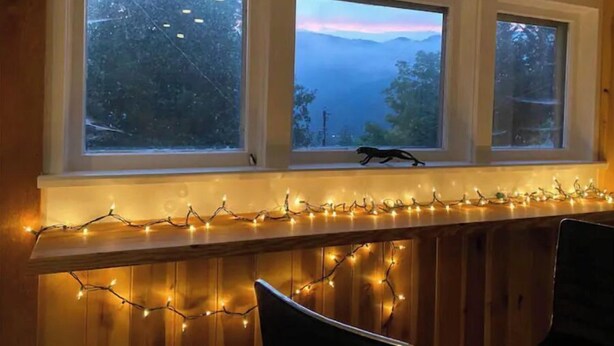 A room with three windows overlooking a forested area, a small table with string lights, and a chair. - Awesome House in Downtown Sylva! (Sylva)