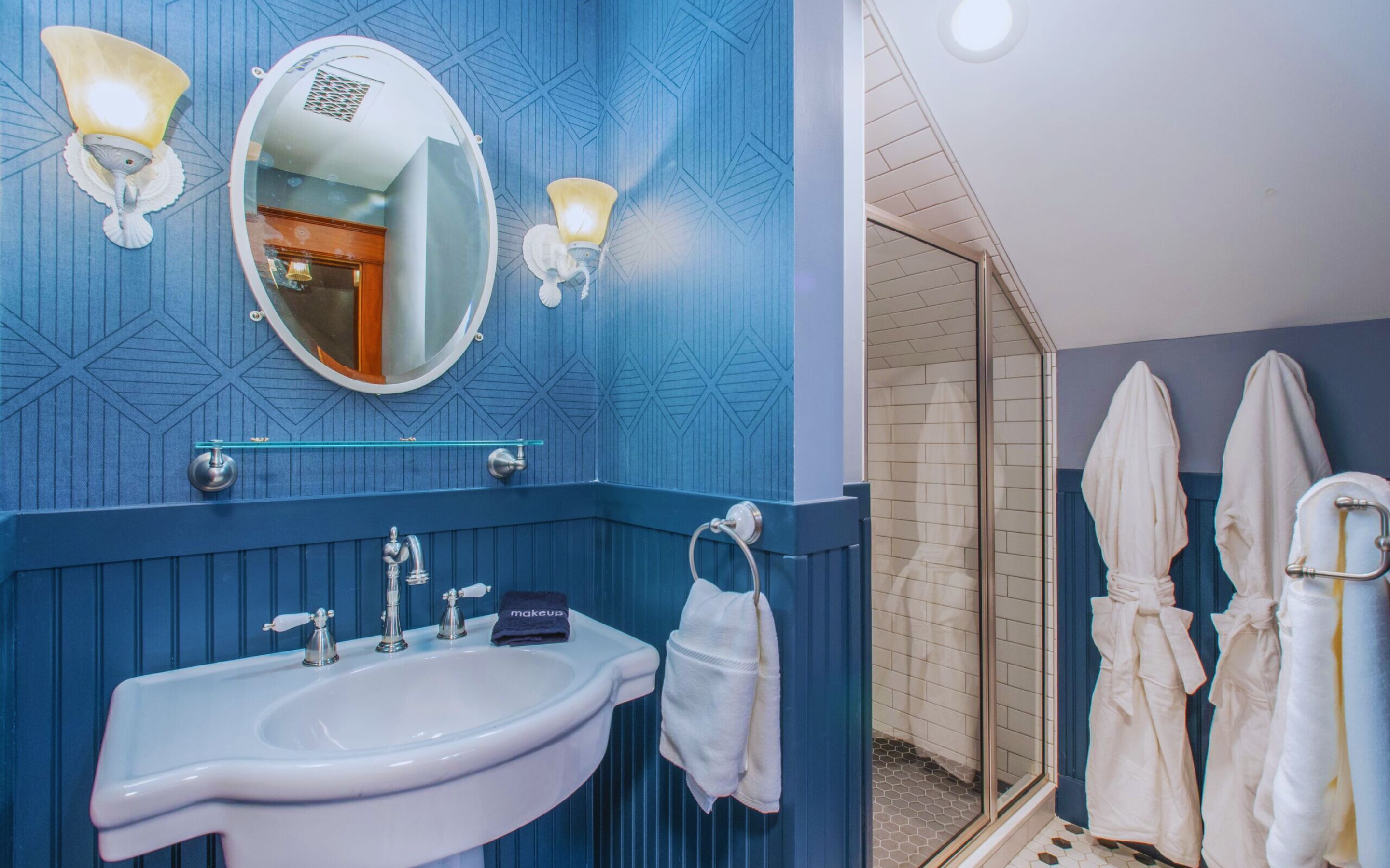 Une salle de bain équipée d’un lavabo, d’un miroir et de deux appliques murales. On y trouve des serviettes et un porte-peignoir avec des peignoirs.
