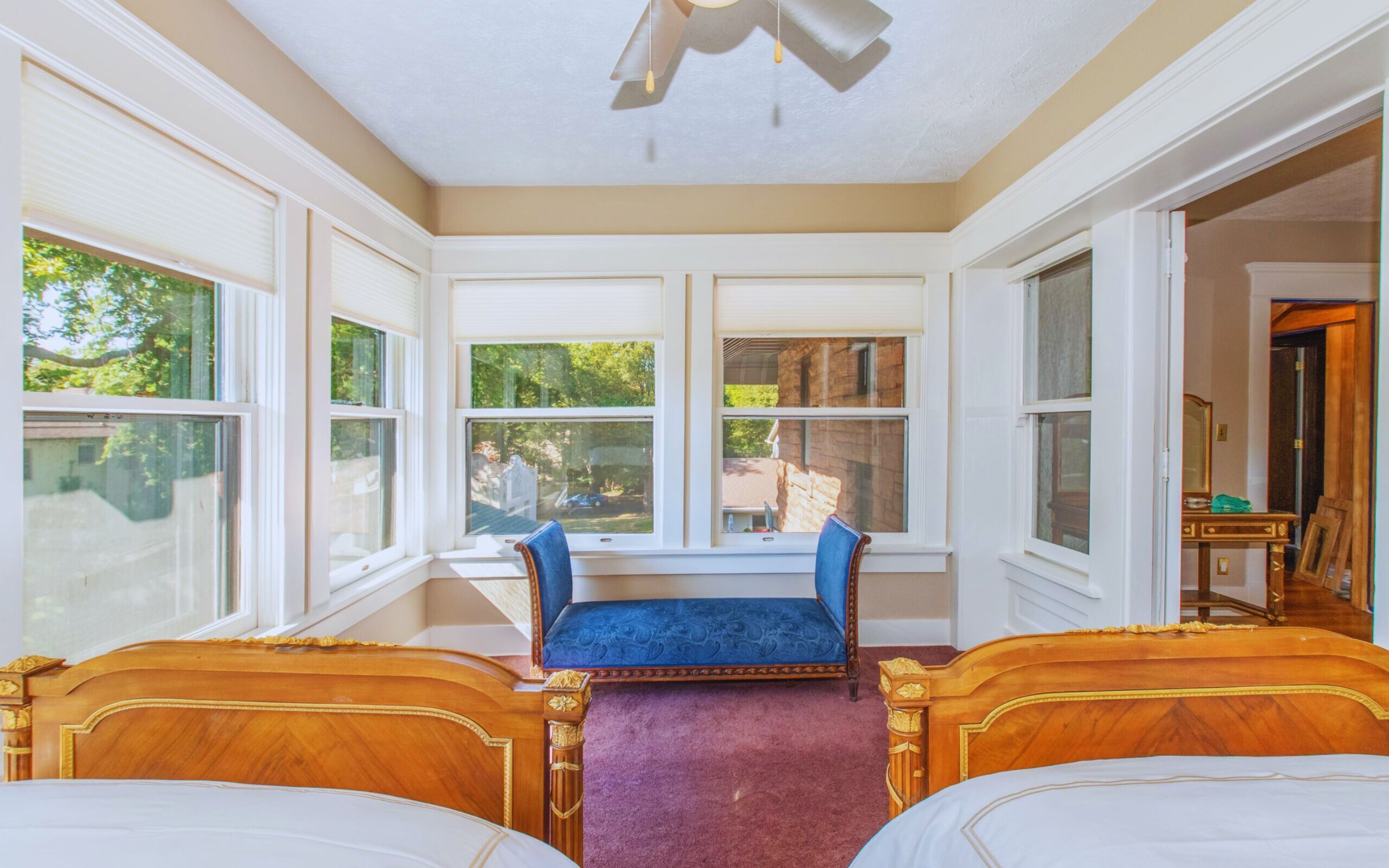 A room with two wooden beds, a blue bench, large windows, and a view of greenery outside.