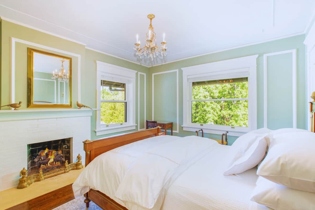A bedroom with a bed, a fireplace, a chandelier, and windows with a view of trees.