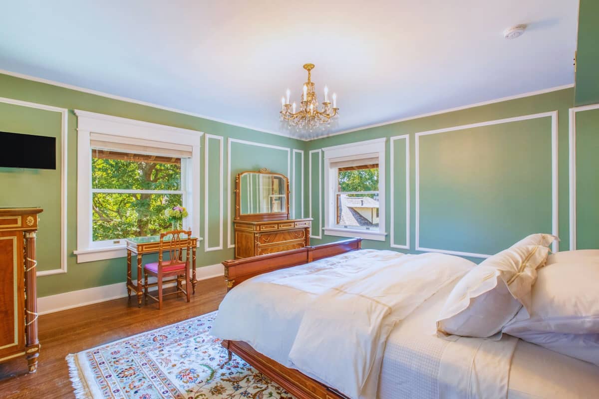 A bedroom with a large bed, a dresser, a chair, and a mirror. There are two windows with views of greenery outside.