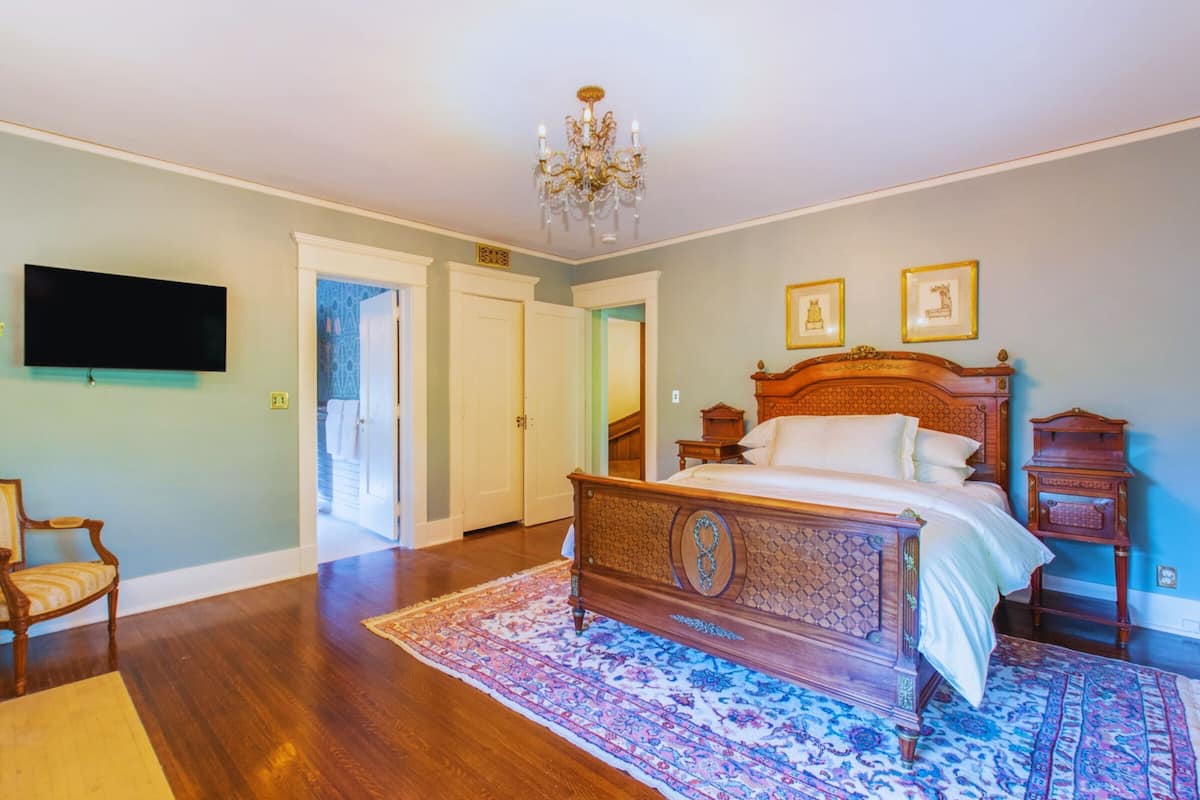 A bedroom with a wooden bed, a chandelier, a television, and a door leading to another room.