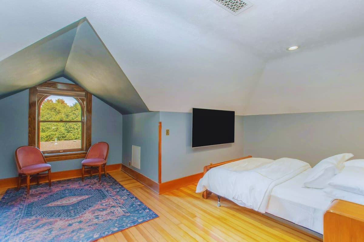 A bedroom with a sloped ceiling, a large window, a TV, a bed, and a chair.