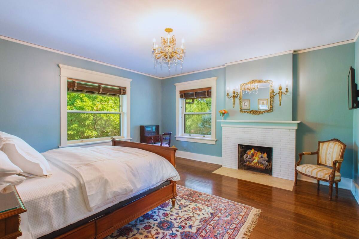 A bedroom with a large bed, two windows, a fireplace, and a chandelier.