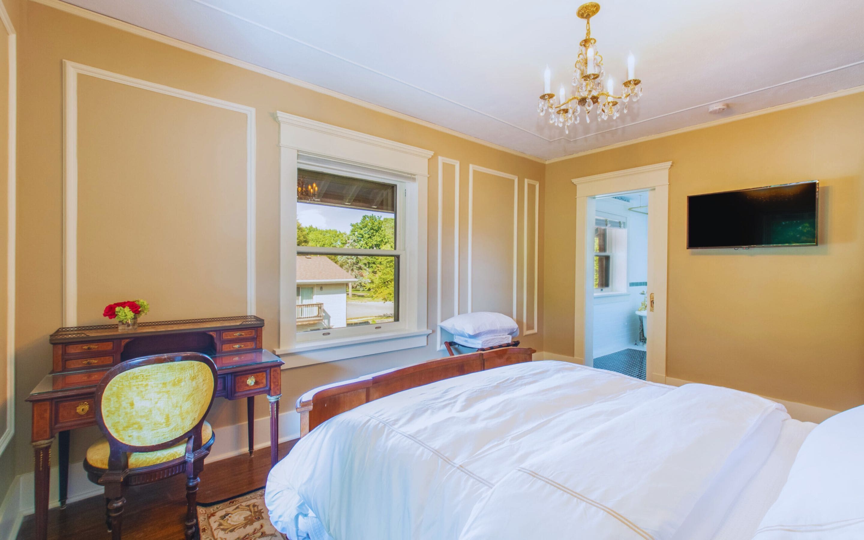 Quarto com cama, escrivaninha com cadeira, televisão e janela com vista para área verde.