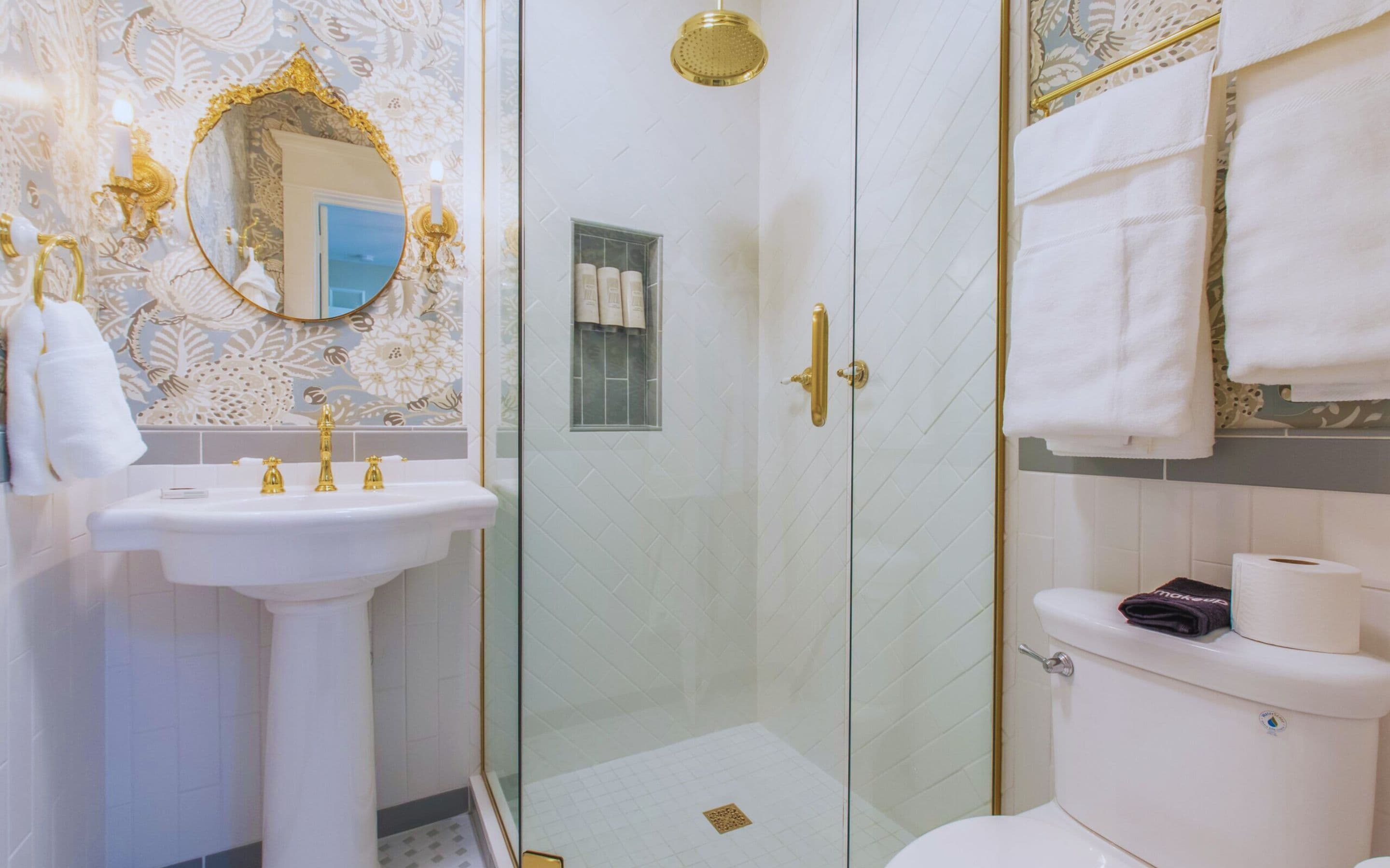 A bathroom with a glass-enclosed shower, a white sink with gold fixtures, and a toilet with a paper towel dispenser.