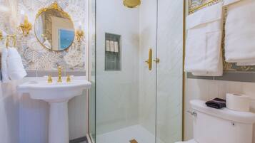 A bathroom with a glass-enclosed shower, a white sink with gold fixtures, and a toilet with a paper towel dispenser.