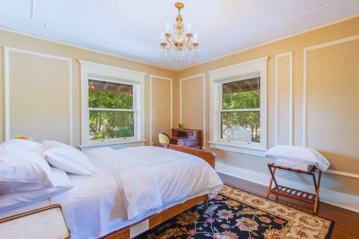 A bedroom with a large bed, two windows, a chandelier, and a small wooden table.