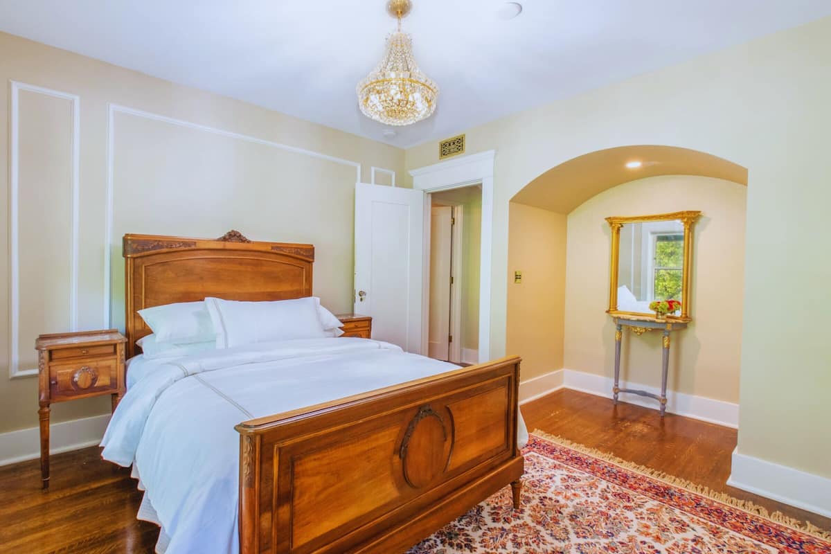 A bedroom with a wooden bed, a nightstand, a dresser, a chandelier, and a mirror with a floral pattern rug.