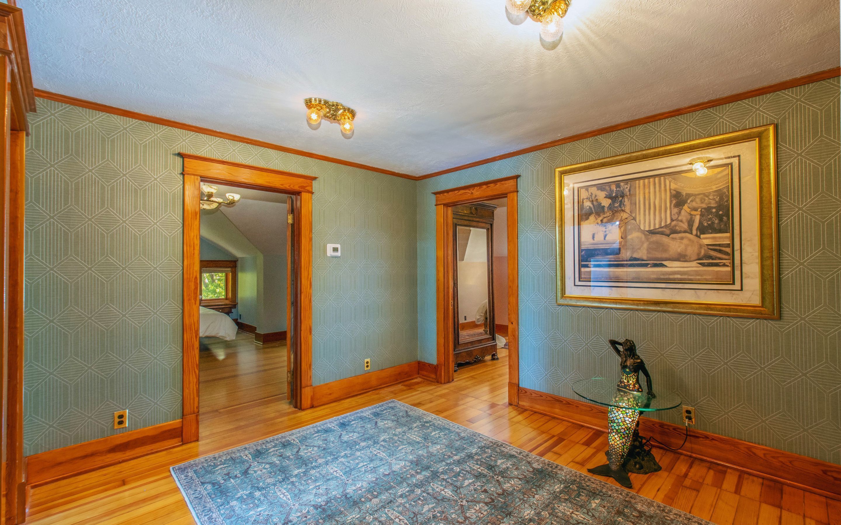 A hallway with patterned wallpaper, wooden trim, a glass table with a decorative base, and a framed picture on the wall.
