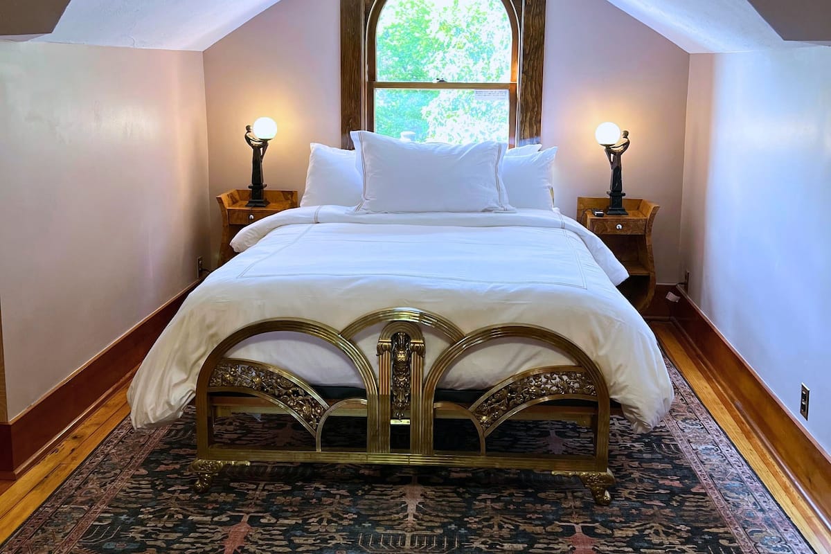 A bedroom with a vaulted ceiling, a large bed with ornate headboard, two bedside lamps, and a window with a view of greenery.