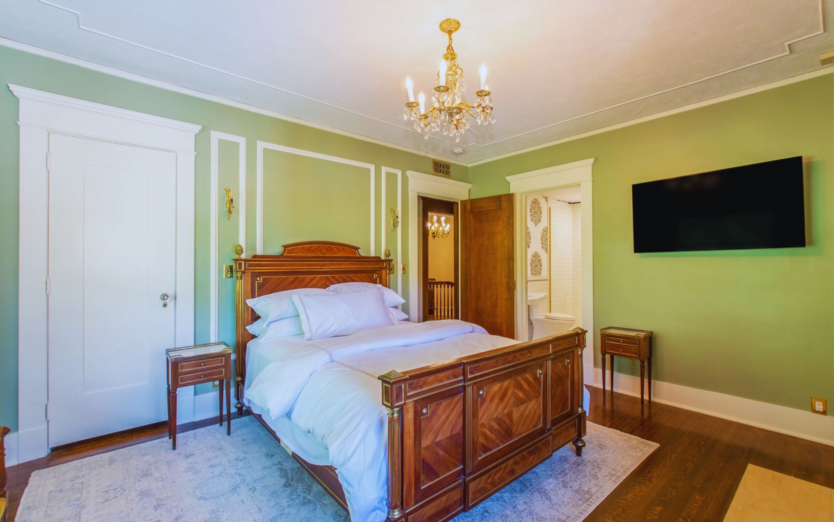 A bedroom with a wooden bed, a chandelier, a television, and a bathroom visible through an open door.