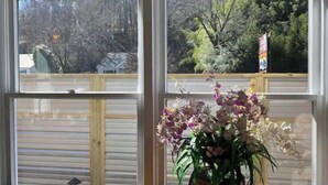 A room with a large window, a wooden chair, and a potted plant with flowers. - Not available (Sylva)