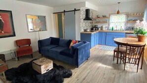 A modern living room with a blue sofa, a dining table with chairs, a kitchen area with blue cabinets, and a black furry rug. - Not available (Sylva)