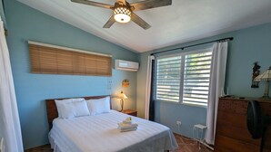 A bedroom with a bed, a ceiling fan, a window with blinds, a nightstand, a dresser, and a lamp. - Cheerful Home with Gorgeous Ocean Views and Pool (Vieques,)