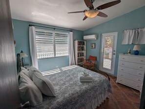 A bedroom with a bed, a ceiling fan, a bookshelf, a chair, a dresser, and a window with blinds.
