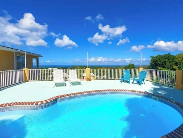 A pool area with a view of the ocean, surrounded by a white fence and blue chairs.