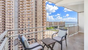 A balcony with wicker chairs and a table, overlooking a high-rise building and a clear sky.