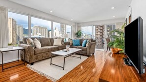 A modern living room with a sofa, coffee table, and TV, large windows with a city view, and a balcony with plants.