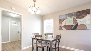 A dining area with a wooden table and chairs, a ceiling light fixture, and a large abstract painting on the wall.