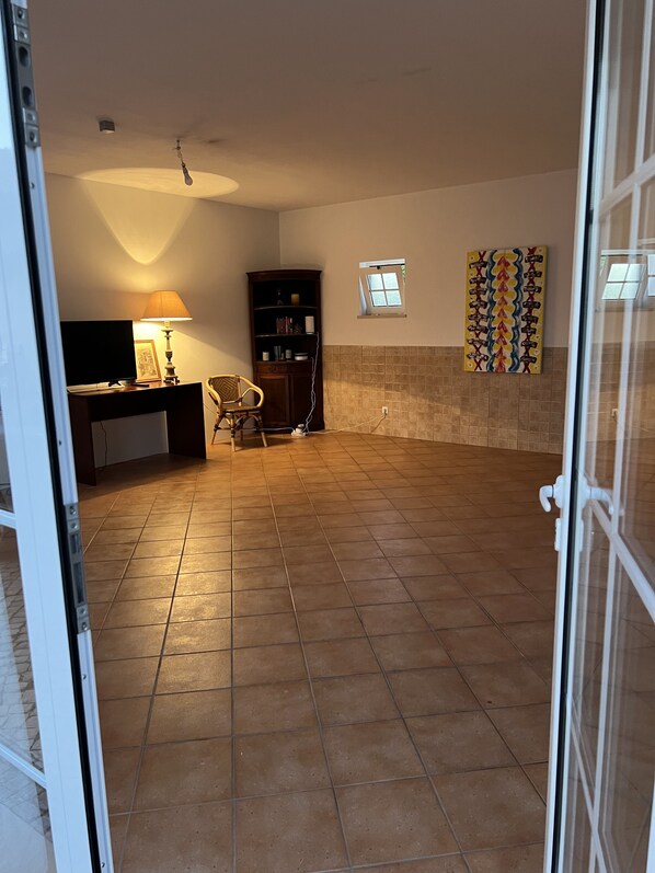 A spacious room with a tiled floor, a desk with a lamp, a bookshelf, a chair, and a window.